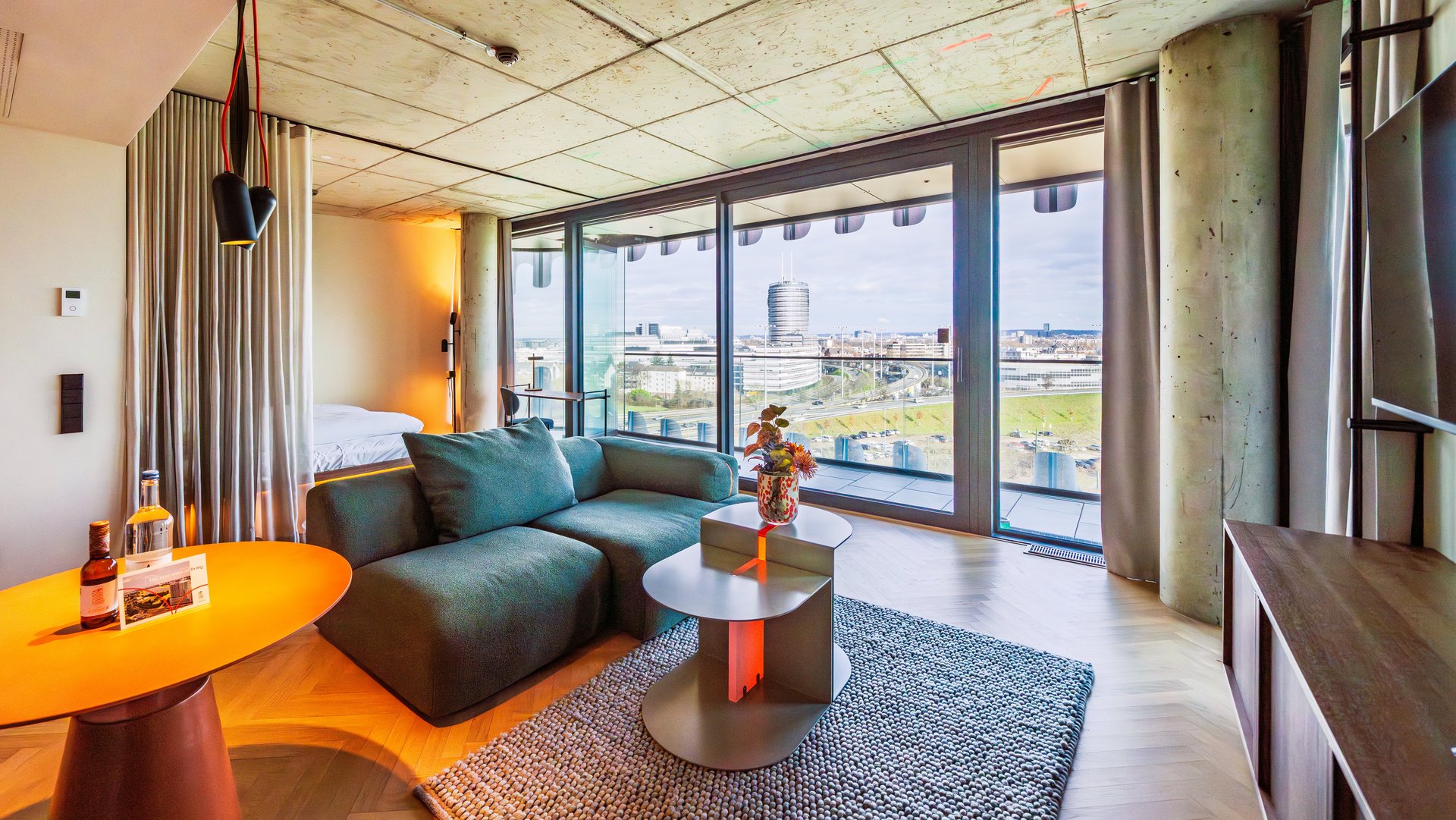 Panorama-Ansicht des Wohnzimmers im The Zipper Hotel mit grünem Ecksofa und Zugang zum Balkon.