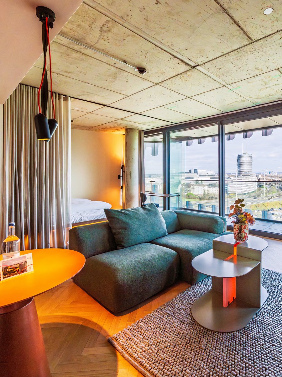 Panorama-Ansicht des Wohnzimmers im The Zipper Hotel mit grünem Ecksofa und Zugang zum Balkon.