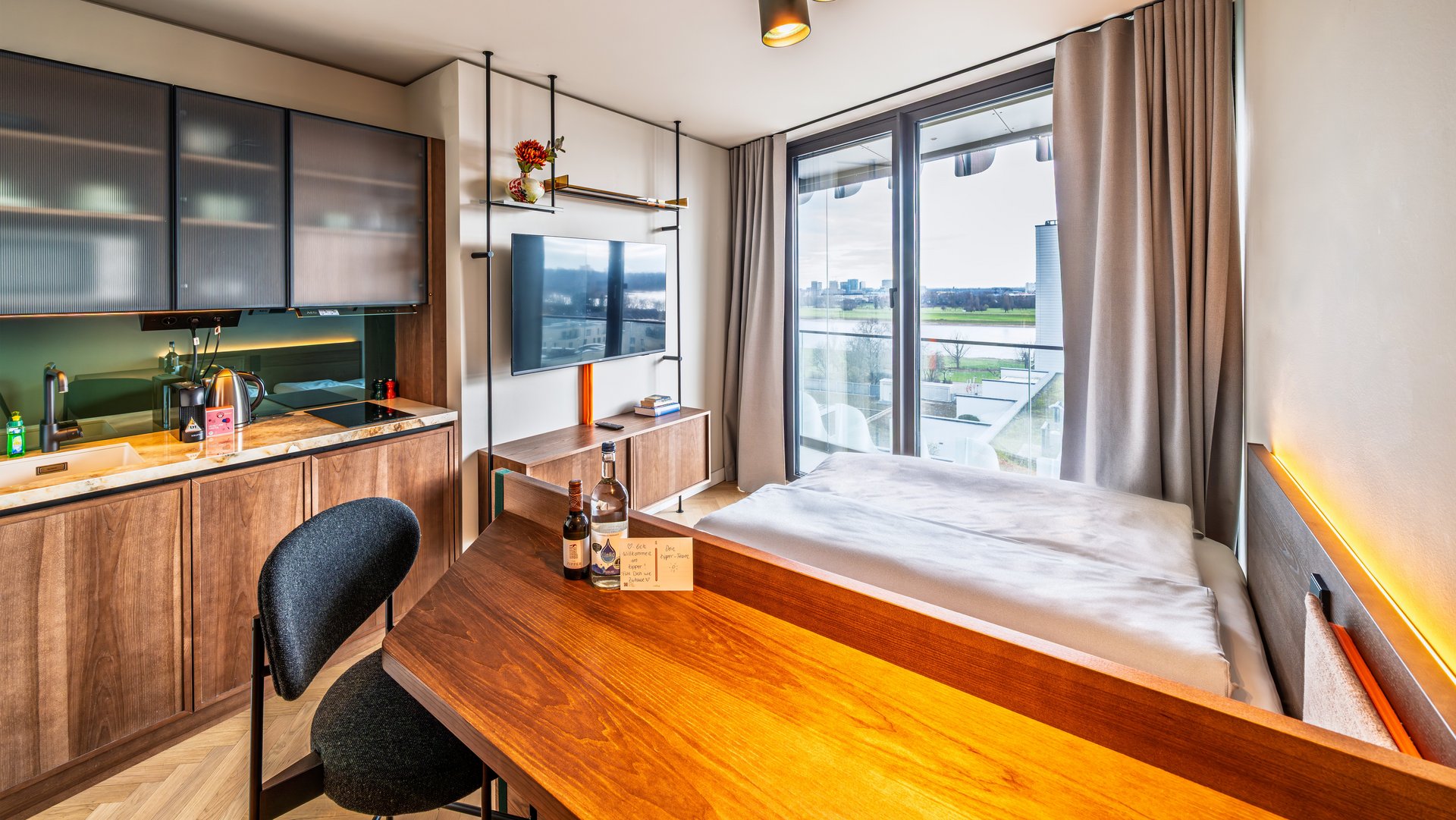 Modernes Apartment im The Zipper Hotel mit Holztisch, Kochnische und Blick auf das Bett sowie ein großes Fenster.