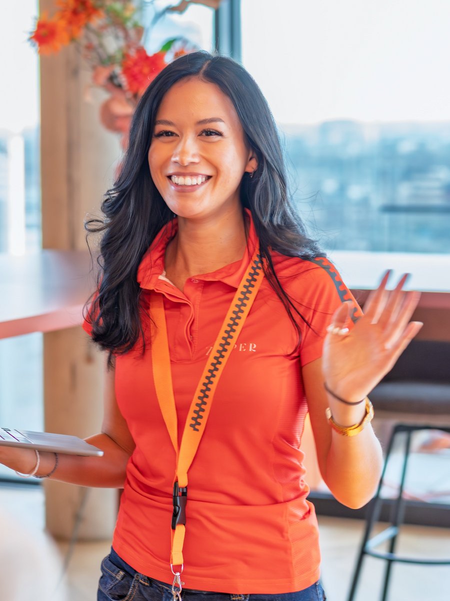 Eine lächelnde Mitarbeiterin in orangefarbenem Poloshirt begrüßt Gäste