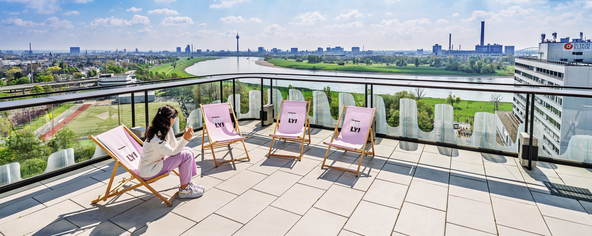 Eine Frau sitzt entspannt in einem Liegestuhl auf der modernen Dachterrasse von The Zipper und genießt den weiten Panorama-Ausblick über den Rhein und Düsseldorf.