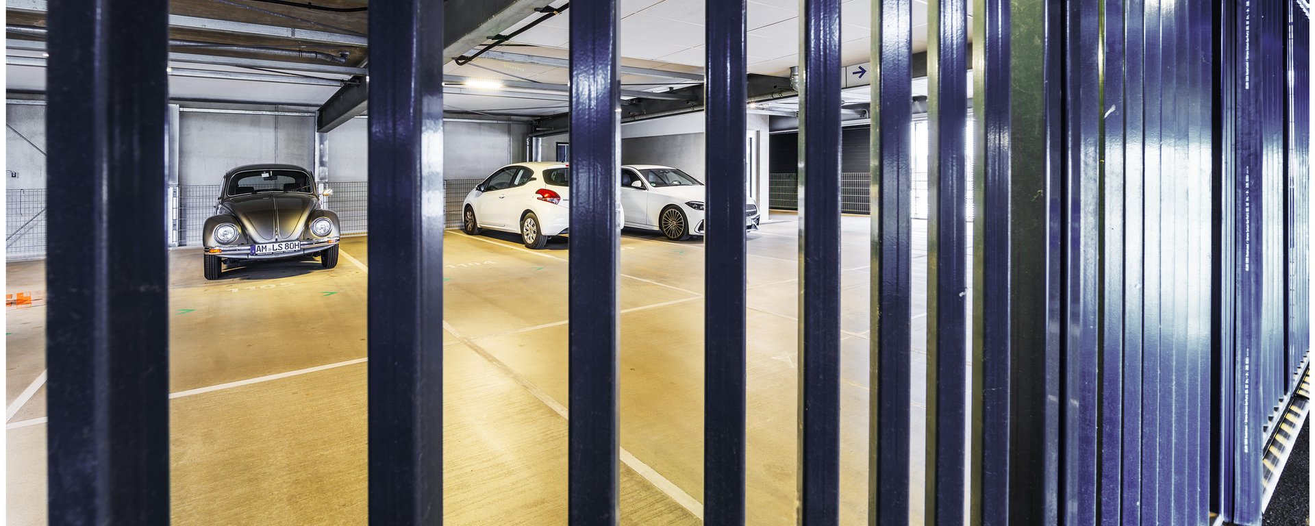 Perspektive durch vertikale, dunkle Metallgitterstäbe hindurch auf die dahinter liegenden, beleuchteten Parkplätze.