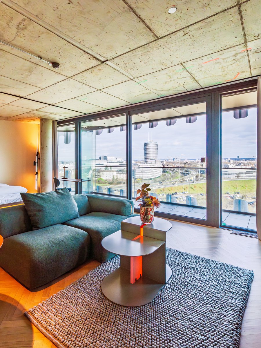 Panorama-Ansicht des Wohnzimmers im The Zipper Hotel mit grünem Ecksofa und Zugang zum Balkon.