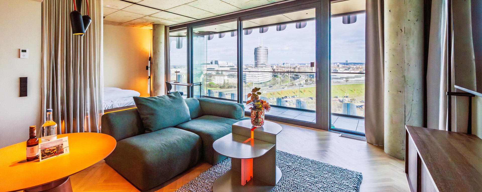 Panorama-Ansicht des Wohnzimmers im The Zipper Hotel mit grünem Ecksofa und Zugang zum Balkon.