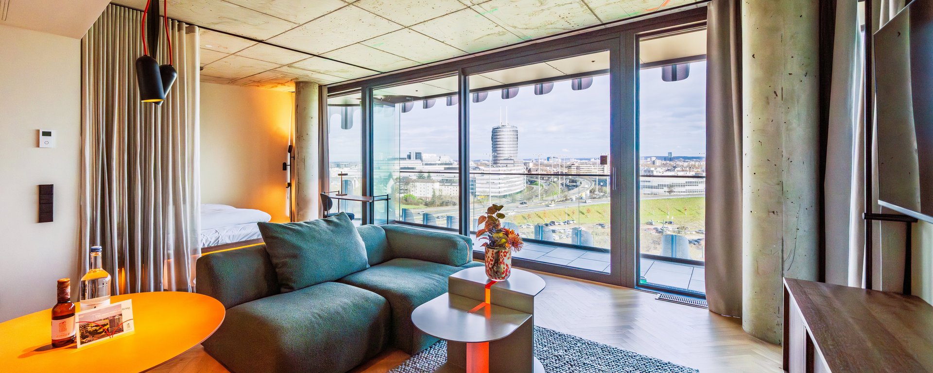 Panorama-Ansicht des Wohnzimmers im The Zipper Hotel mit grünem Ecksofa und Zugang zum Balkon.