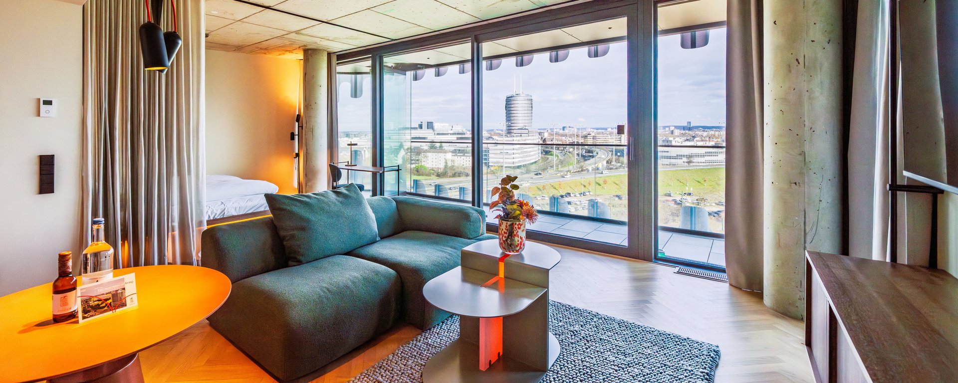 Panorama-Ansicht des Wohnzimmers im The Zipper Hotel mit grünem Ecksofa und Zugang zum Balkon.