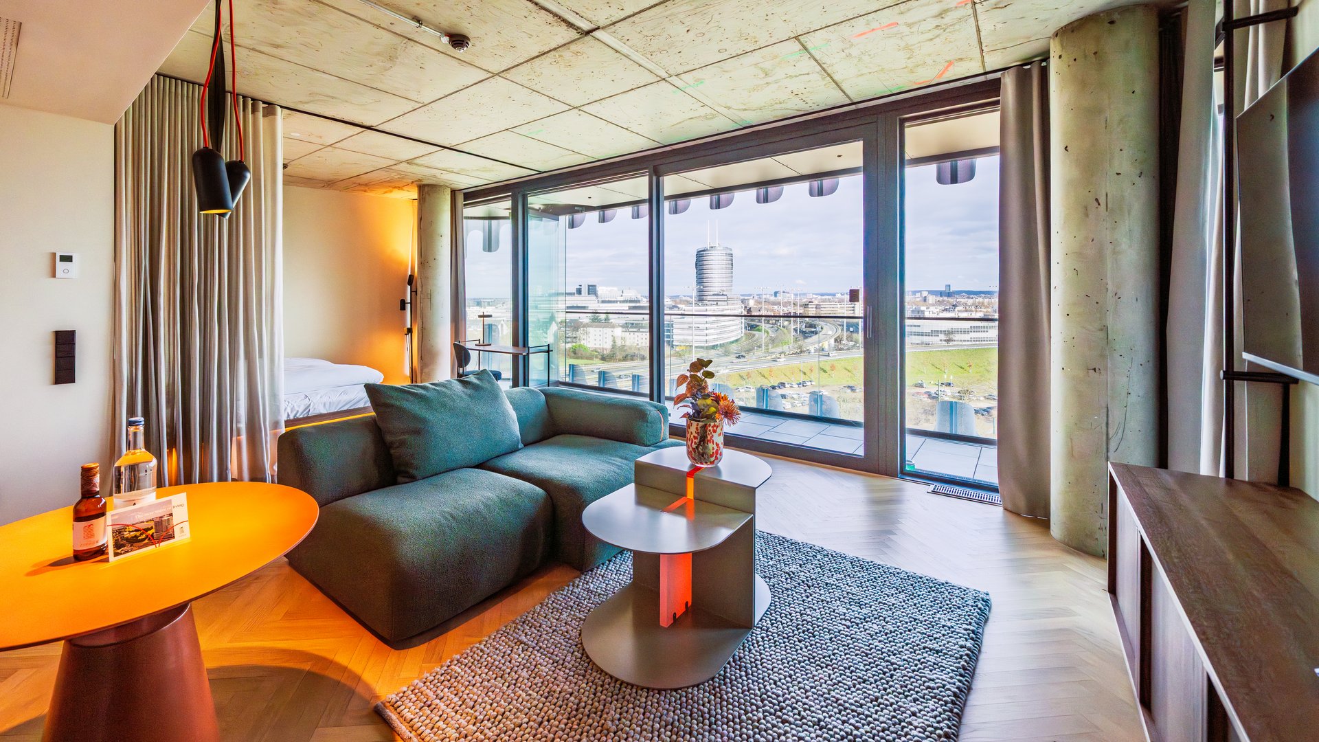 Panorama-Ansicht des Wohnzimmers im The Zipper Hotel mit grünem Ecksofa und Zugang zum Balkon.