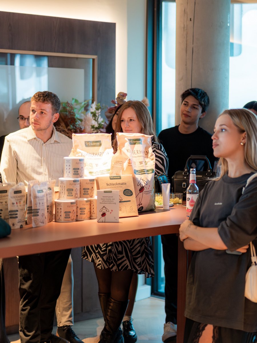Hunderunde-Vortrag im The Zipper Hotel & Apartments Düsseldorf mit Produktpräsentation. Gäste hören beim Hunderunde-Vortrag im The Zipper Hotel & Apartments Düsseldorf aufmerksam zu, Produkte von Hunderunde im Vordergrund.