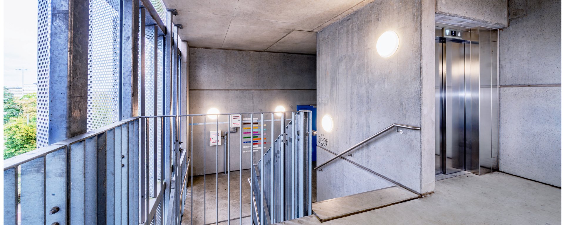 Helles, modernes Treppenhaus im Parkhaus mit einer Beton- und Stahltreppe, Edelstahlgeländern und einem Aufzug.