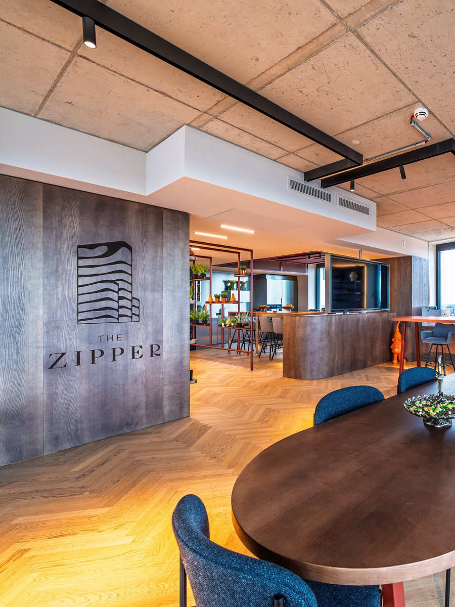 Großzügiger Community Space im The Zipper mit großem Holztisch, blauen Stühlen und modernem Fischgrät-Parkett.