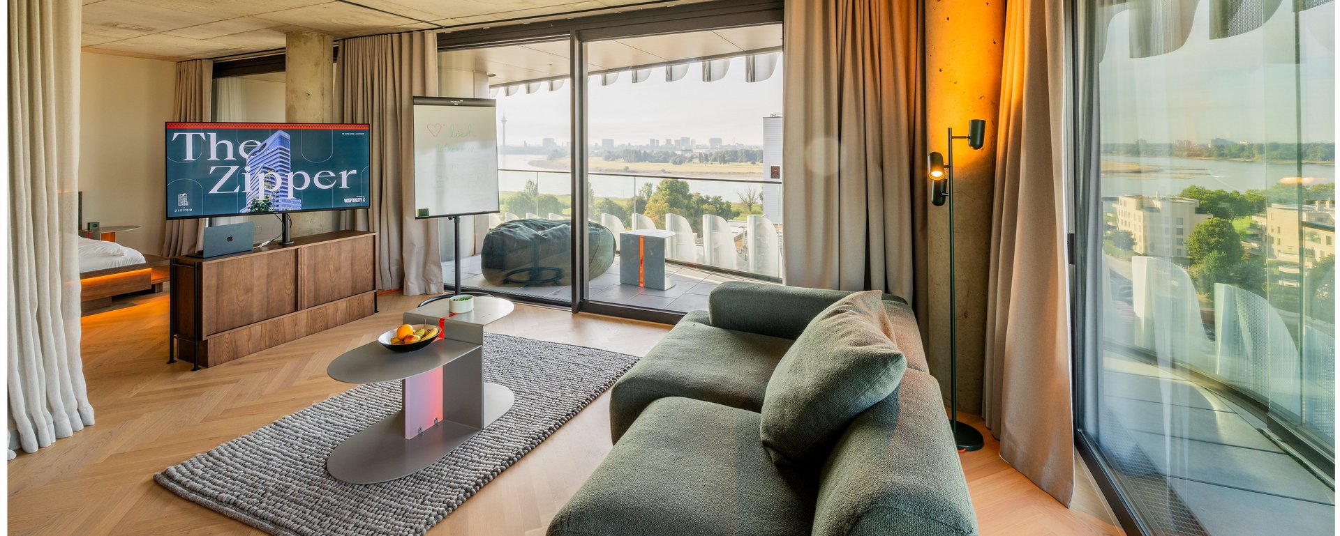 Helles Wohnzimmer einer Hotelsuite mit Sofa, Couchtisch und großem Panoramafenster im THE ZIPPER Hotel Düsseldorf.