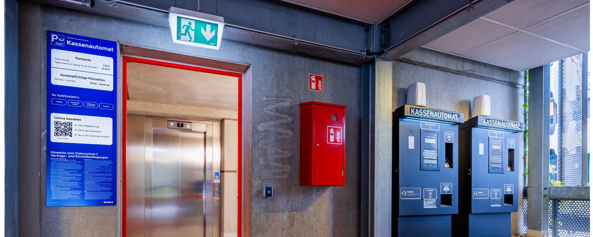 Ein moderner Aufzugsvorraum im Parkhaus mit signalrotem Fußboden, einer grauen Brandschutztür und blauen Hinweisschildern an der Wand.Ein moderner Aufzugsvorraum im Parkhaus mit signalrotem Fußboden, einer grauen Brandschutztür und blauen Hinweisschildern an der Wand.