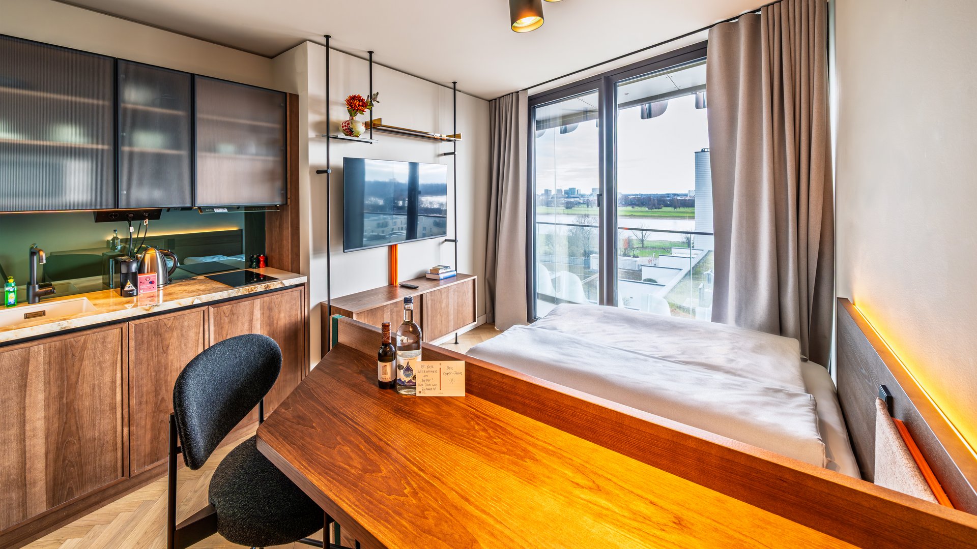 Modernes Apartment im The Zipper Hotel mit Holztisch, Kochnische und Blick auf das Bett sowie ein großes Fenster.