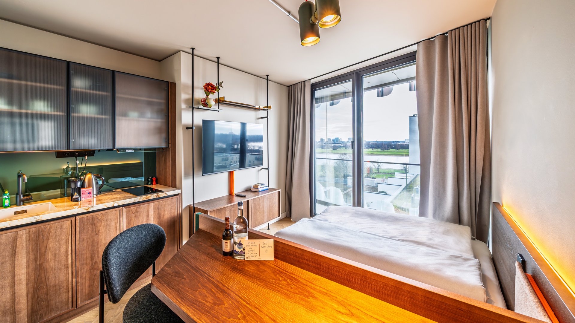 Modernes Apartment im The Zipper Hotel mit Holztisch, Kochnische und Blick auf das Bett sowie ein großes Fenster.