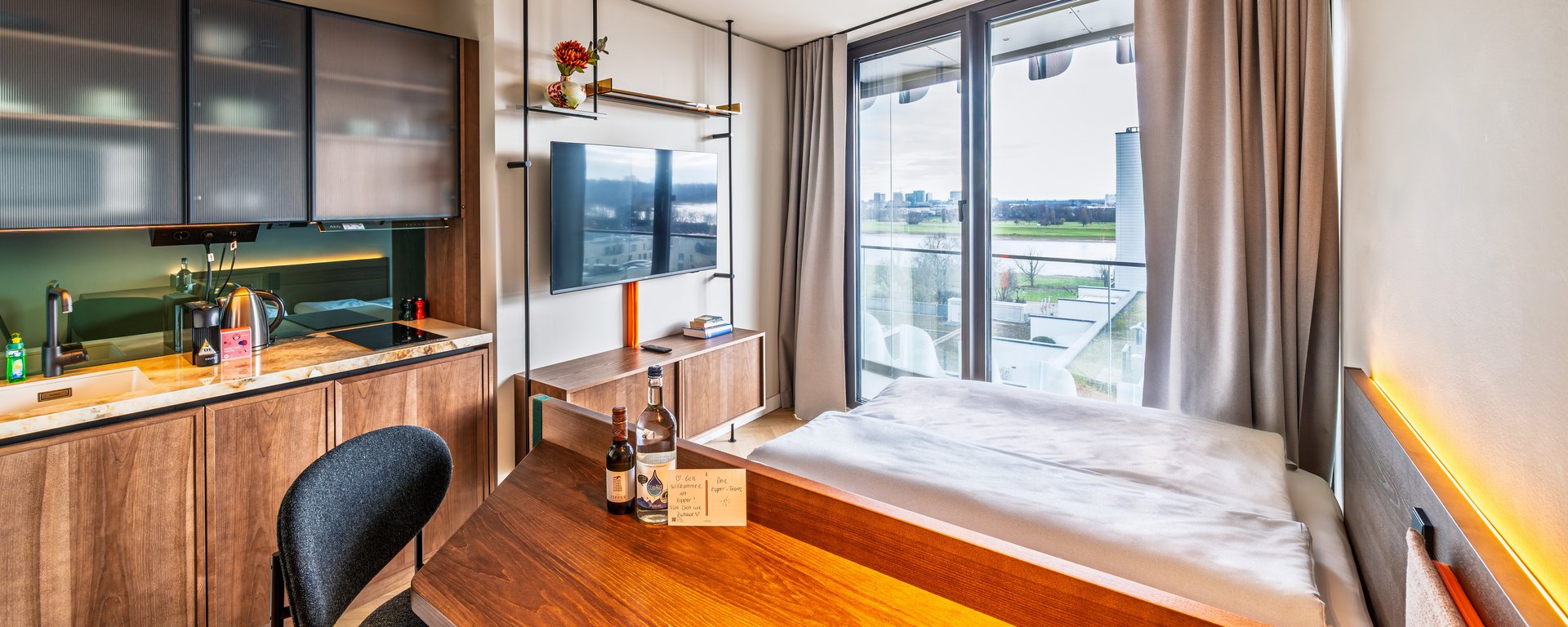 Modernes Apartment im The Zipper Hotel mit Holztisch, Kochnische und Blick auf das Bett sowie ein großes Fenster.