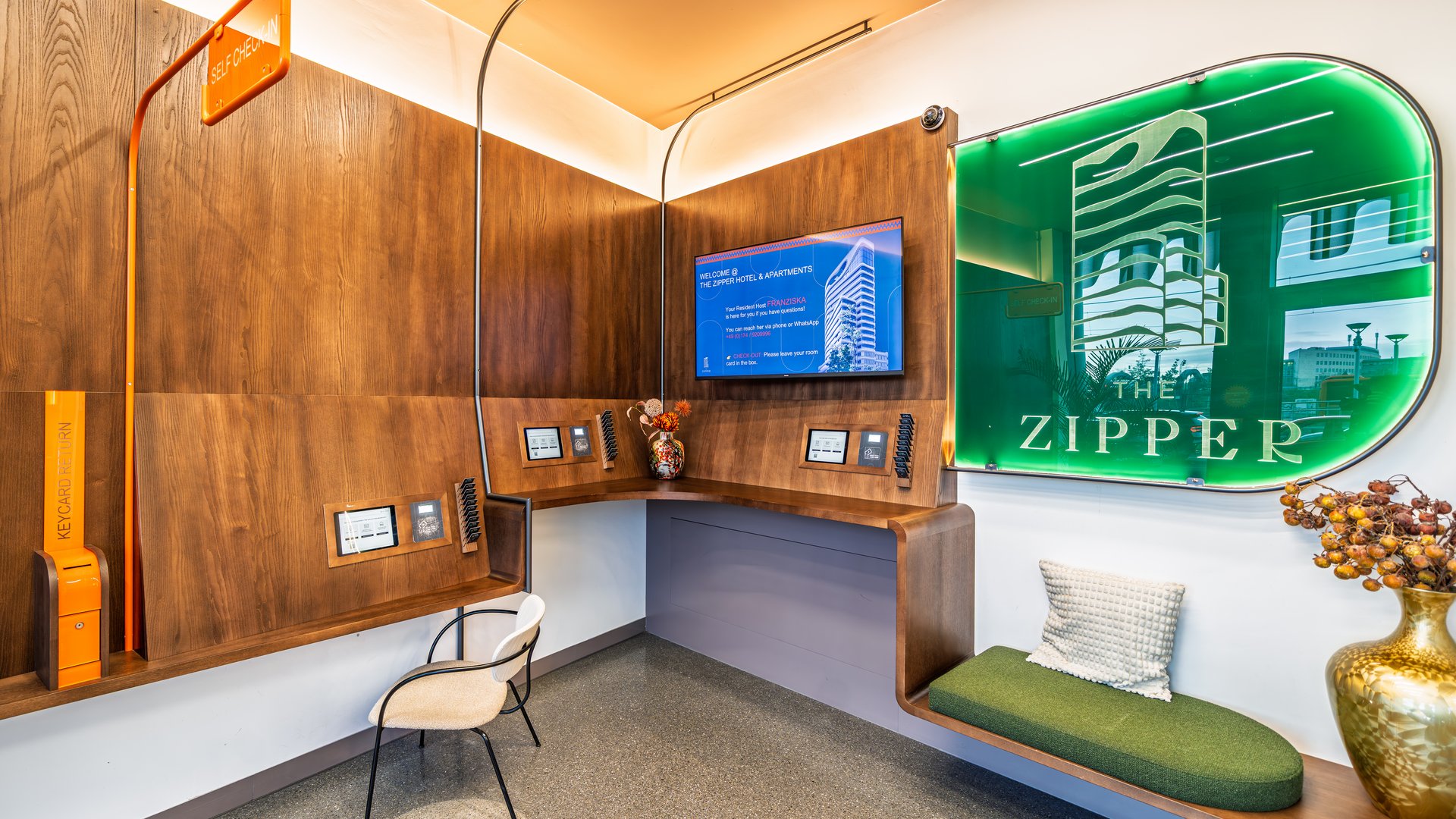 Stylischer Empfangsbereich mit beleuchtetem „The Zipper“ Logo, TV-Screen und Sitzgelegenheiten aus Holz.